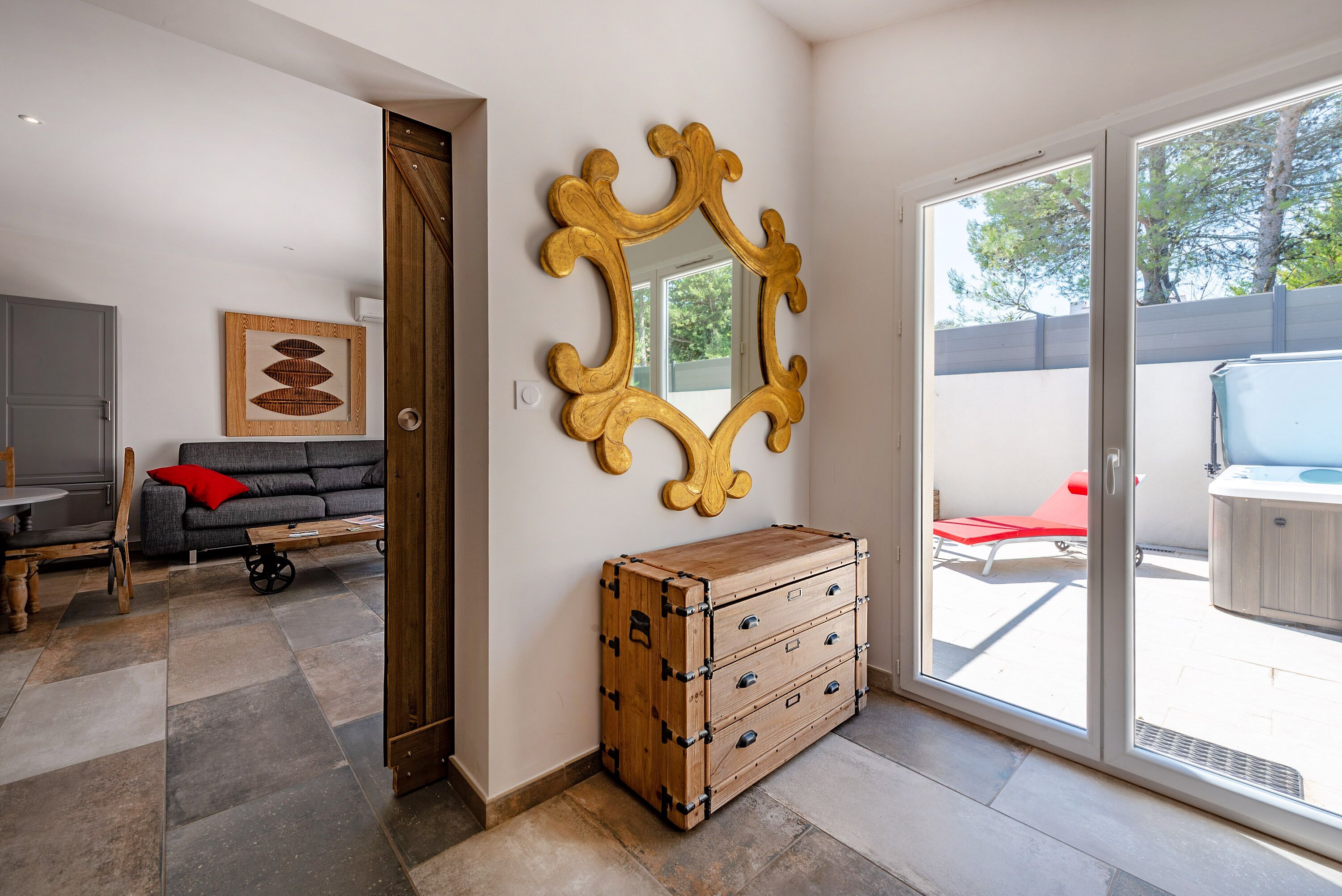 Una sala de estar moderna con un amplio ventanal corredero que da a un área exterior, un aparador de madera, un sofá gris con un cojín rojo y una obra de arte enmarcada en la pared.