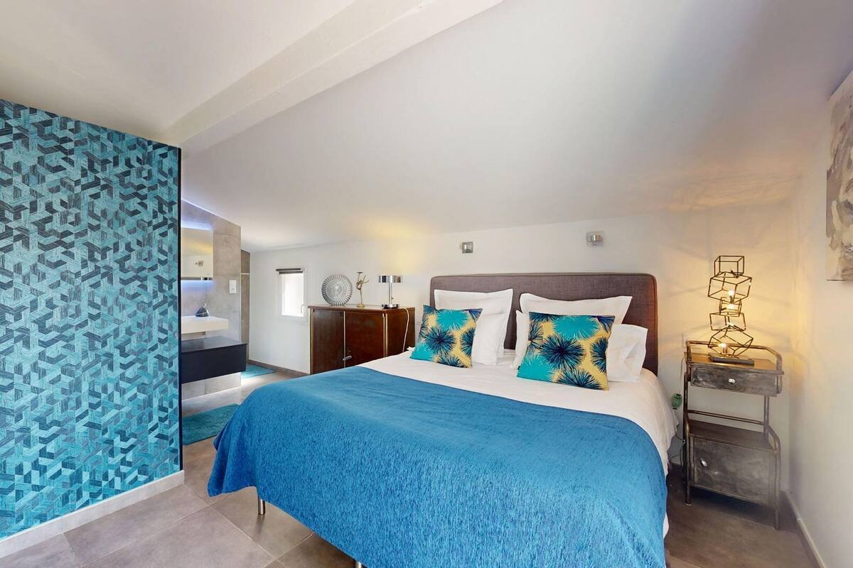Une chambre d’hôtel moderne avec un grand lit, une table de chevet, un miroir et une salle de bain visible à travers une paroi en verre.