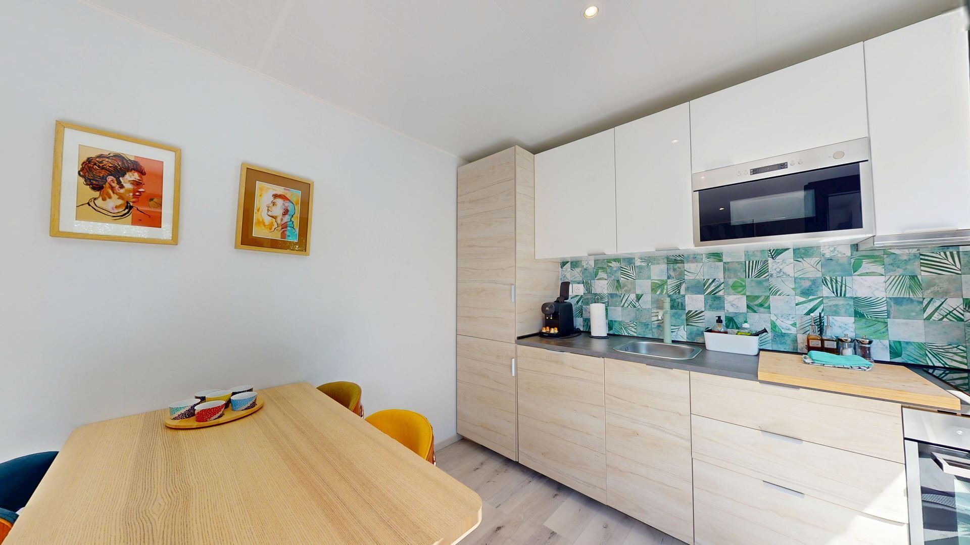 A modern kitchen with a microwave, a coffee machine, and a tiled backsplash. There is a dining table with chairs and two framed artworks on the wall.
