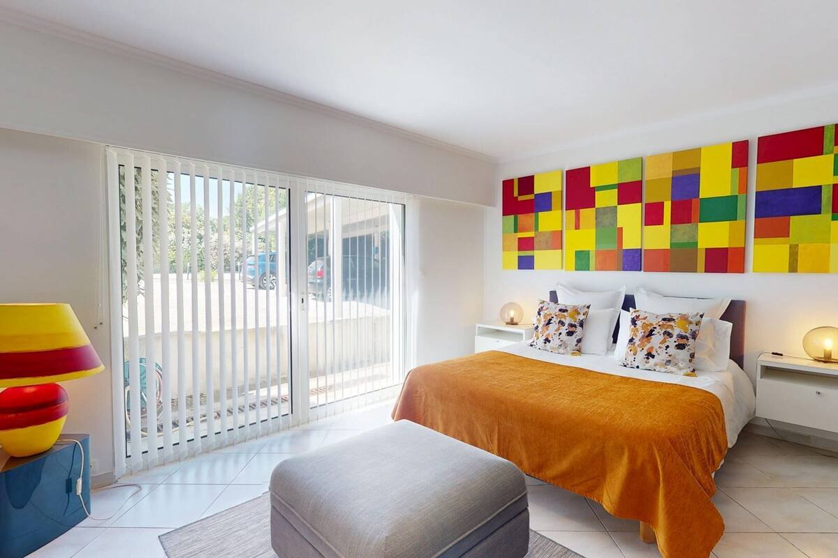 Une chambre à coucher avec un lit, une table de chevet, une lampe, un tableau abstrait aux couleurs vives et une vue à travers une porte coulissante.