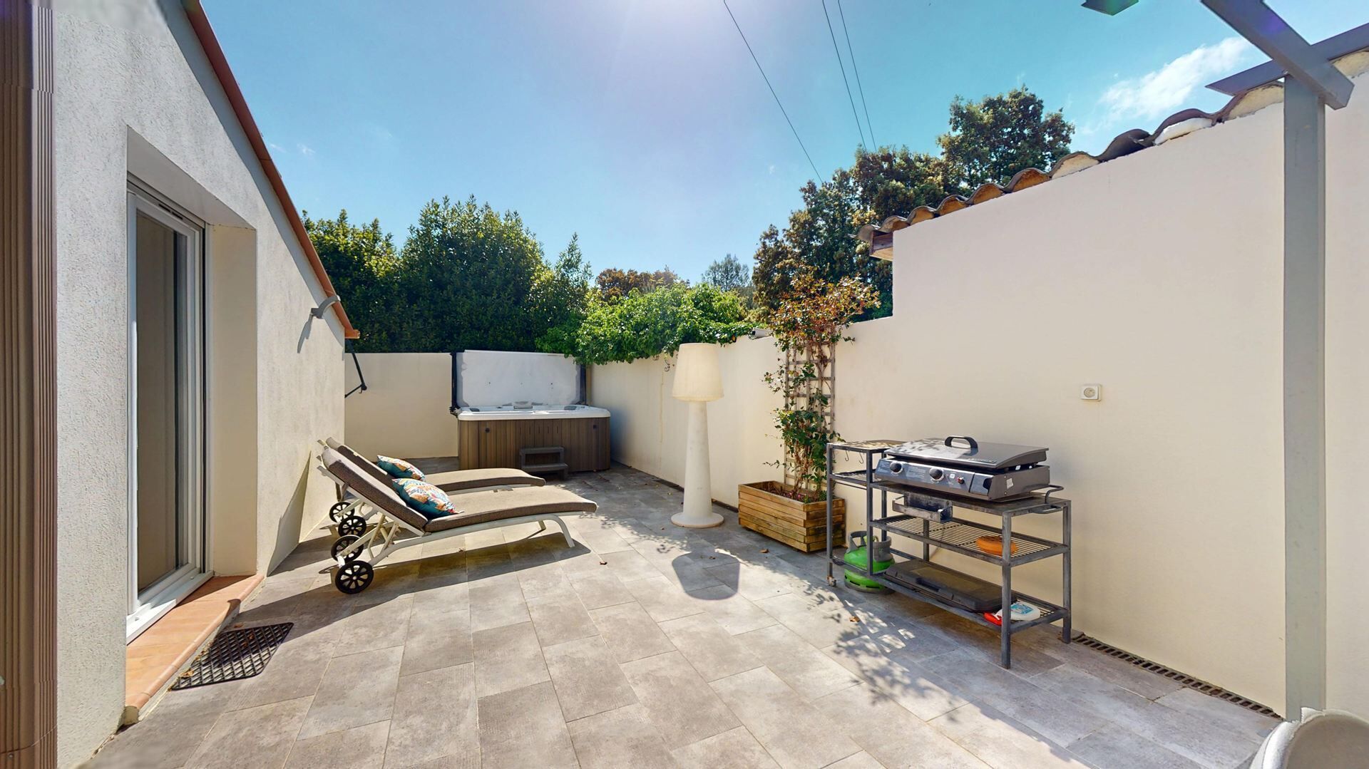 A patio with a hot tub, lounge chairs, a grill, and a small tree.