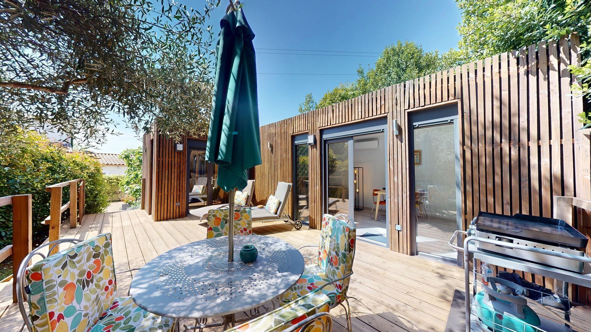 A wooden deck with a round table and chairs, a covered seating area, and a sliding glass door leading to an interior space.