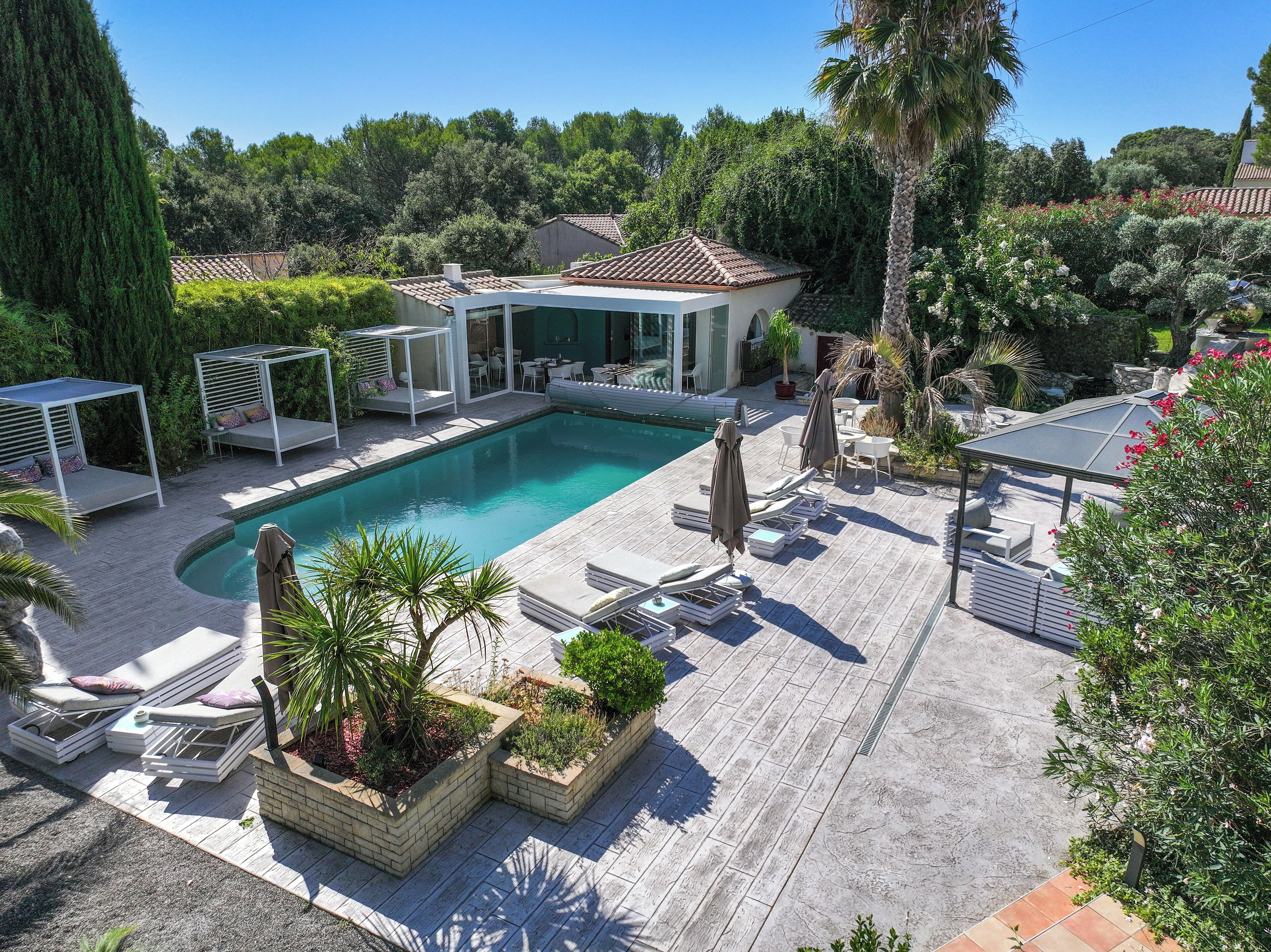 Zona de piscina con sillones de descanso y una pérgola, rodeada de vegetación y un edificio con tejado de tejas.