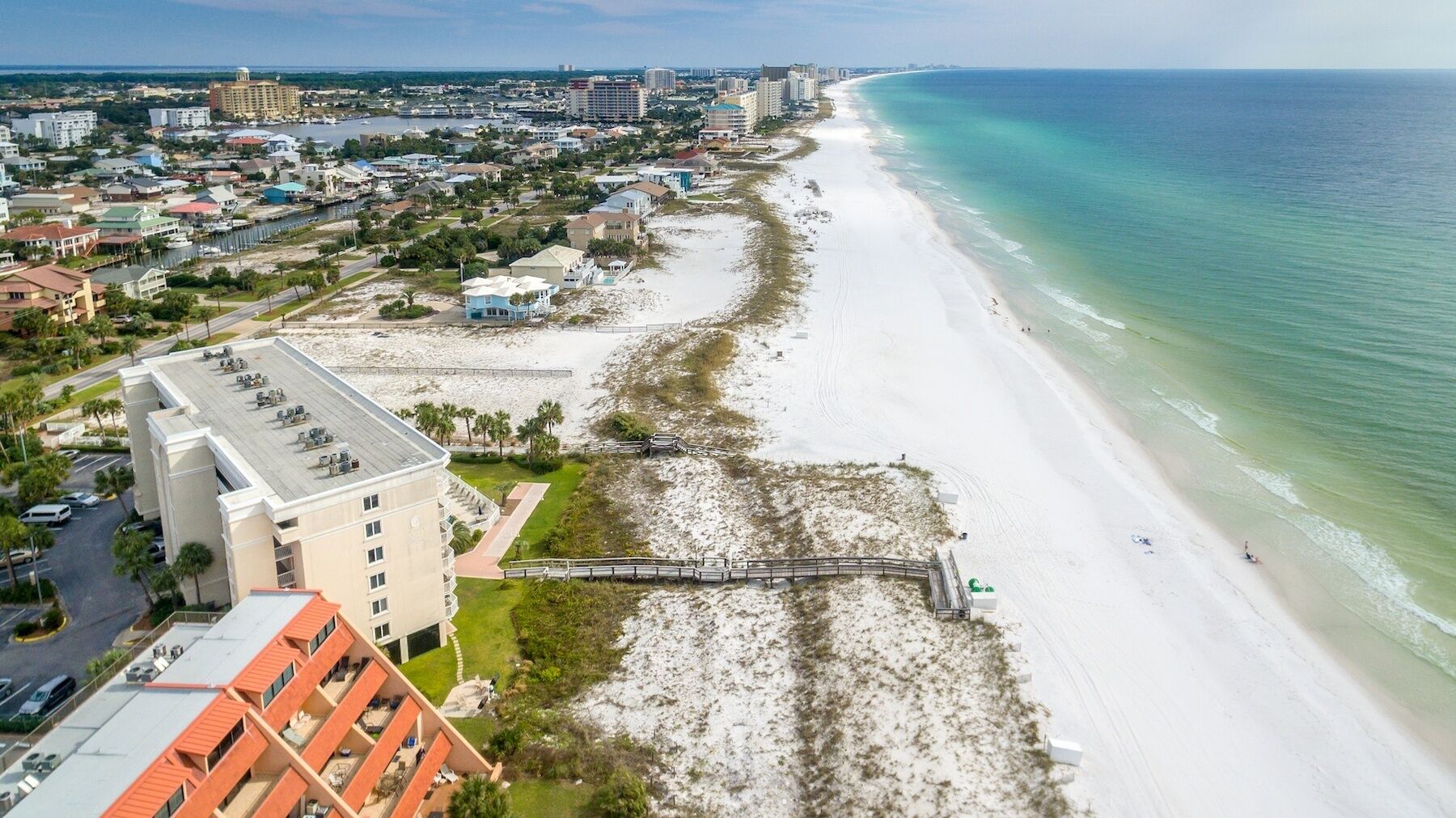 An aerial view of a coastal area with a beach, a pier, and a hotel with multiple floors and balconies.