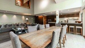 Uma sala de estar ampla com uma grande mesa de jantar de madeira, sofás estofados cinza e uma cozinha ao fundo.