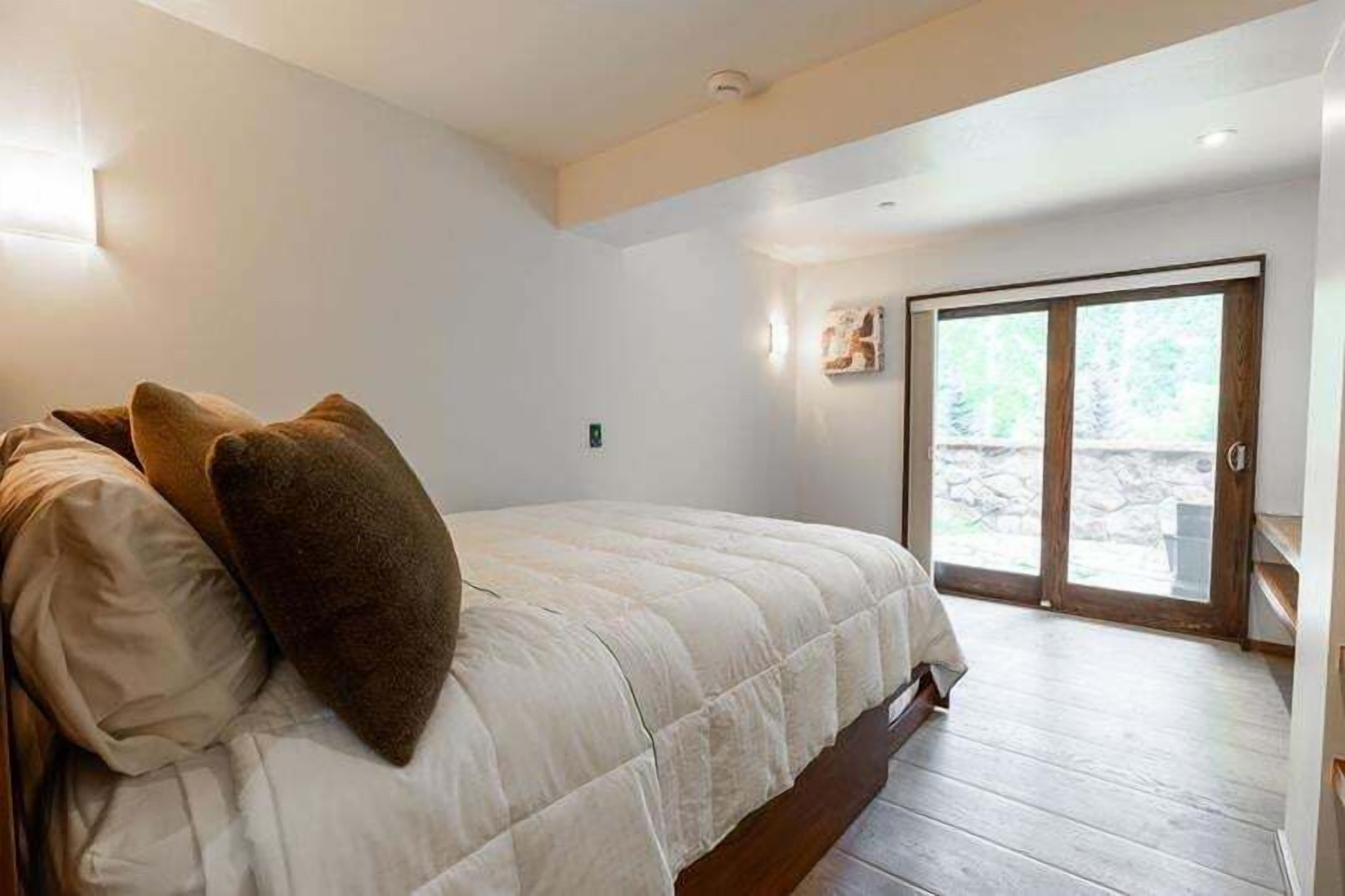 Un dormitorio con cama, almohadas, suelo de madera, puerta corrediza de vidrio y una pequeña lámpara de pared.
