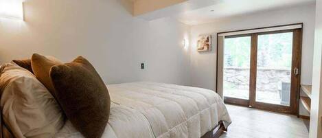 Un dormitorio con cama, almohadas, suelo de madera, puerta corrediza de vidrio y una pequeña lámpara de pared.