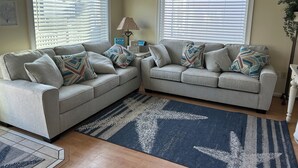 Un salon avec deux canapés gris, des coussins décoratifs à motifs, un tapis bleu et blanc et une horloge au mur.