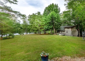 A cabin by a lake with a grassy yard and a large tree in the foreground.