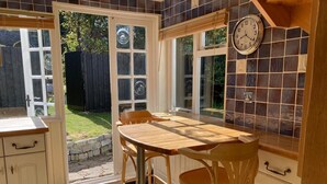 Une cuisine avec une table et des chaises en bois, une horloge au mur et une vue sur le jardin grâce à de grandes fenêtres.