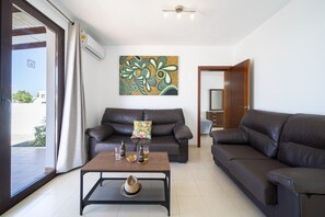 A living room with two sofas, a coffee table with bottles and glasses, a painting on the wall, and a door leading to a bathroom.