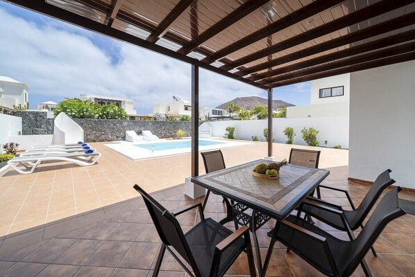 A covered outdoor area with a pool, lounge chairs, and a table with fruit.