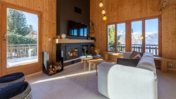Un intérieur de chalet chaleureux avec une cheminée, des murs en bois, un canapé et de grandes fenêtres offrant une vue sur les montagnes.