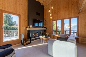 A cozy cabin interior with a fireplace, wooden walls, a sofa, and large windows offering mountain views. - Chalet La Chenevière (Haute-Nendaz)