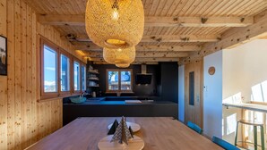Un intérieur en bois avec une longue table à manger, des lampes en osier suspendues et un coin cuisine équipé d’un téléviseur.