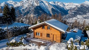 Une cabane en bois, dont le toit est recouvert de neige, entourée d’arbres et de montagnes également enneigés, qui se dressent à l’arrière-plan.