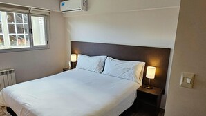 A hotel room with a large bed, two bedside tables with lamps, a window with blinds, and an air conditioning unit. - Hotel Bait (General Roca)