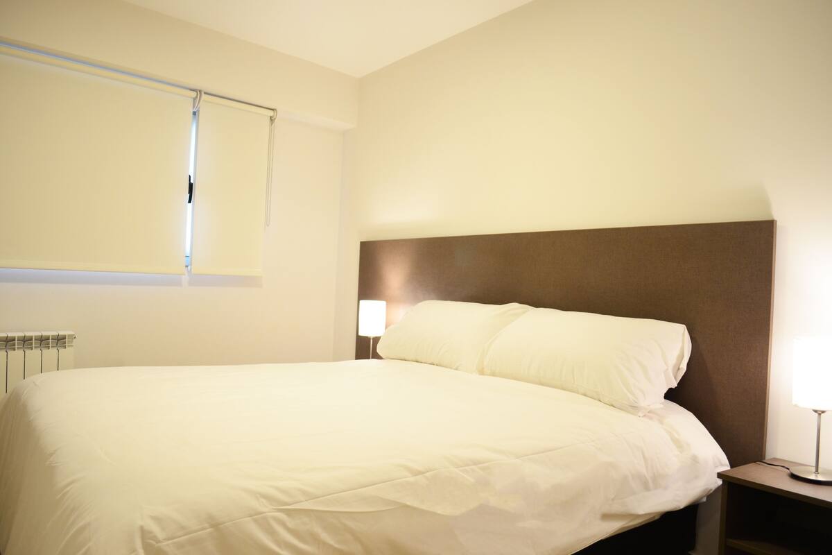 Une chambre d’hôtel avec un grand lit, une table de chevet avec une lampe et une fenêtre avec des stores.