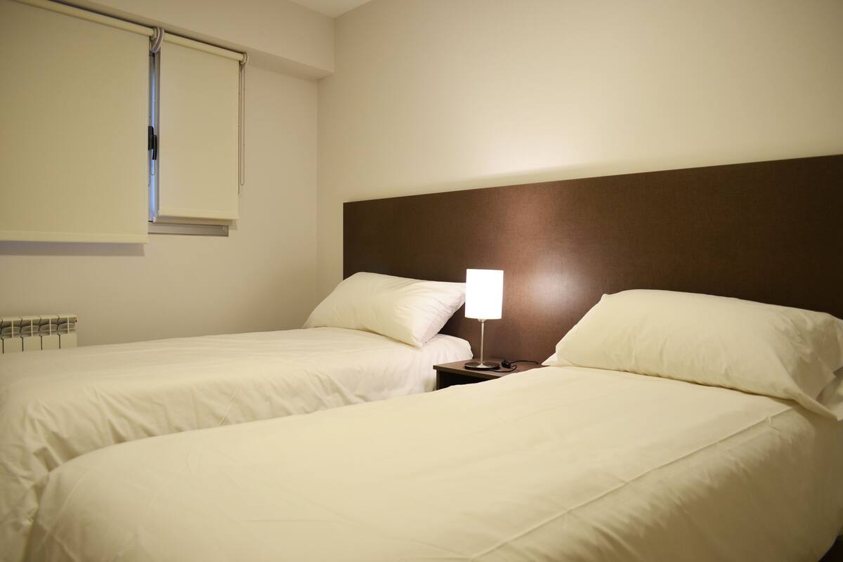 Une chambre d’hôtel avec deux lits, une table de chevet avec une lampe, une fenêtre avec des stores et un radiateur.
