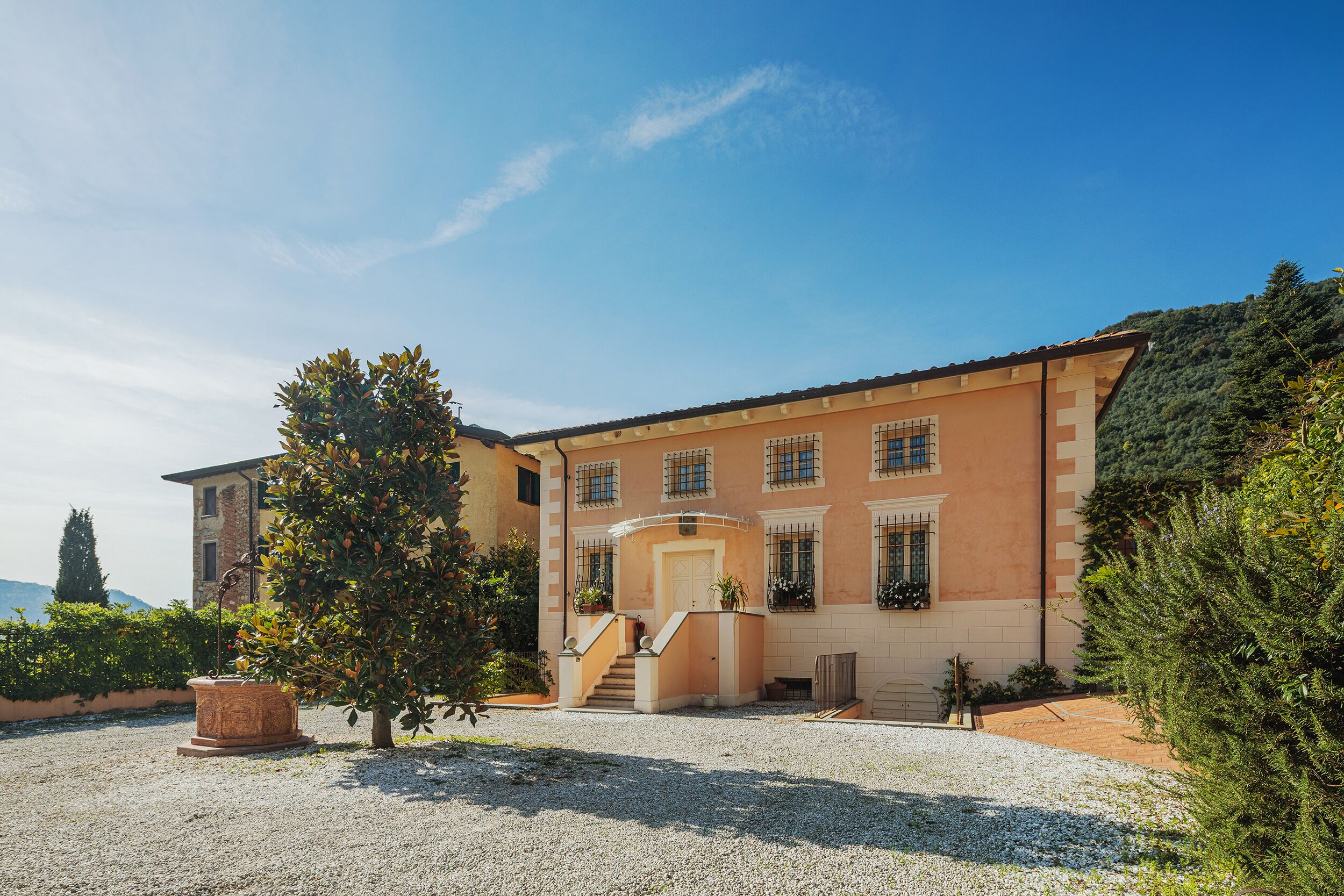 A two-story building with a central entrance, a gravel driveway, and a garden with a large tree.