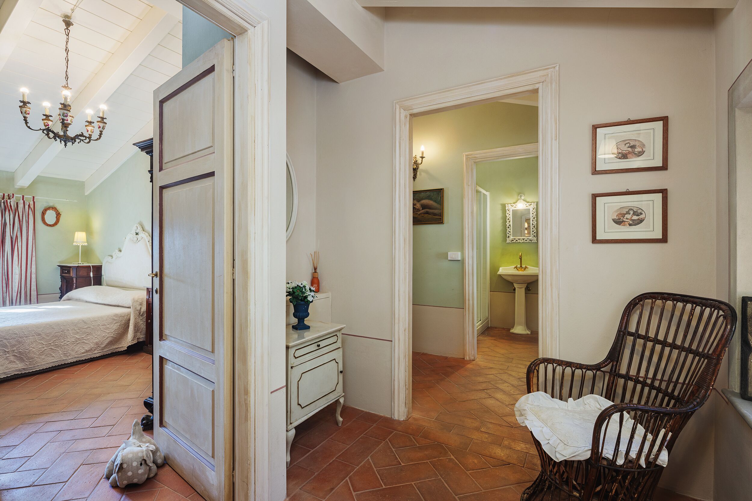 A bedroom with a bed, a dresser, and a chair. There is a hallway leading to another room with a sink and a mirror.