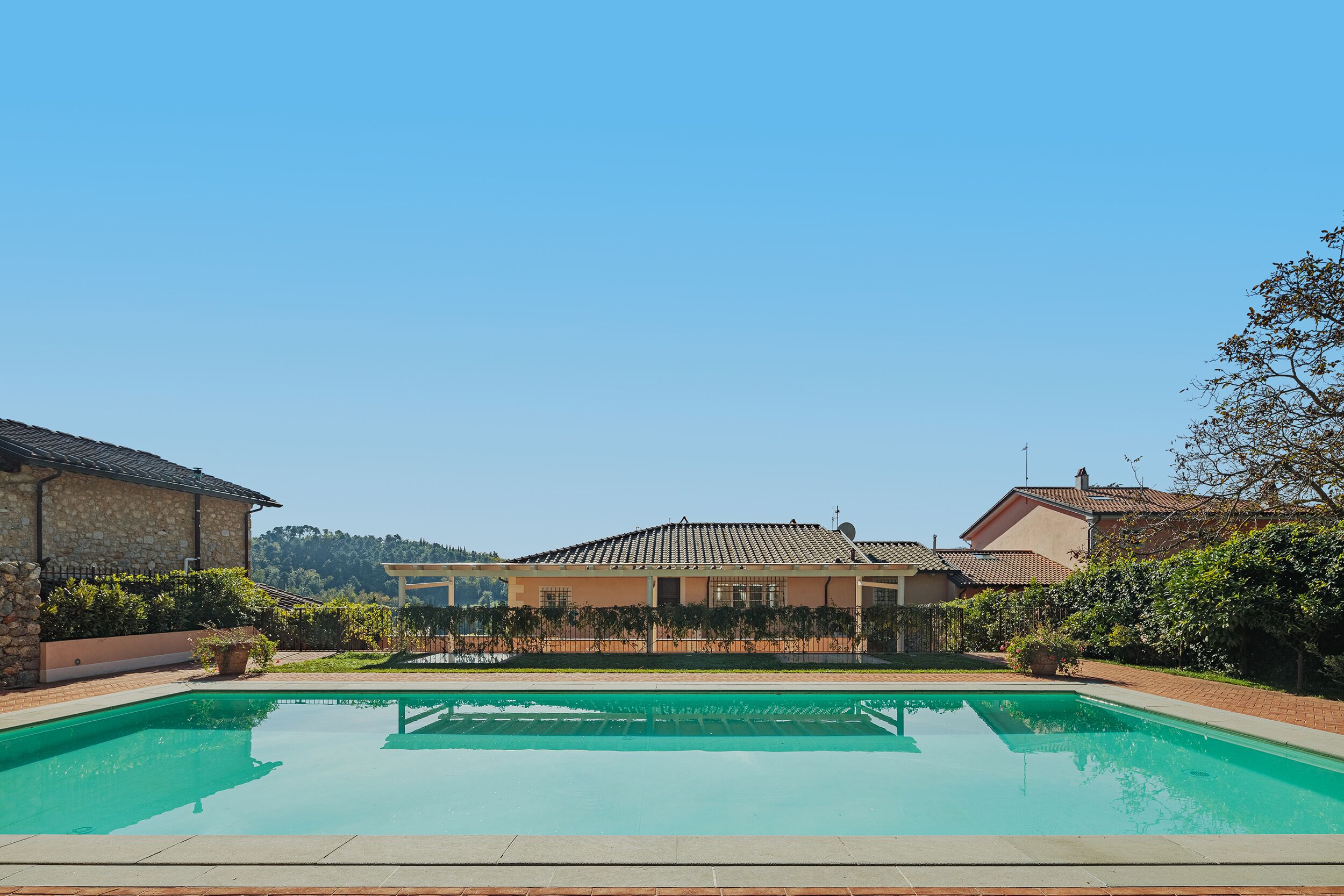Una piscina con un cielo azul despejado al fondo, rodeada por un patio de ladrillo y una casa con techo de tejas.