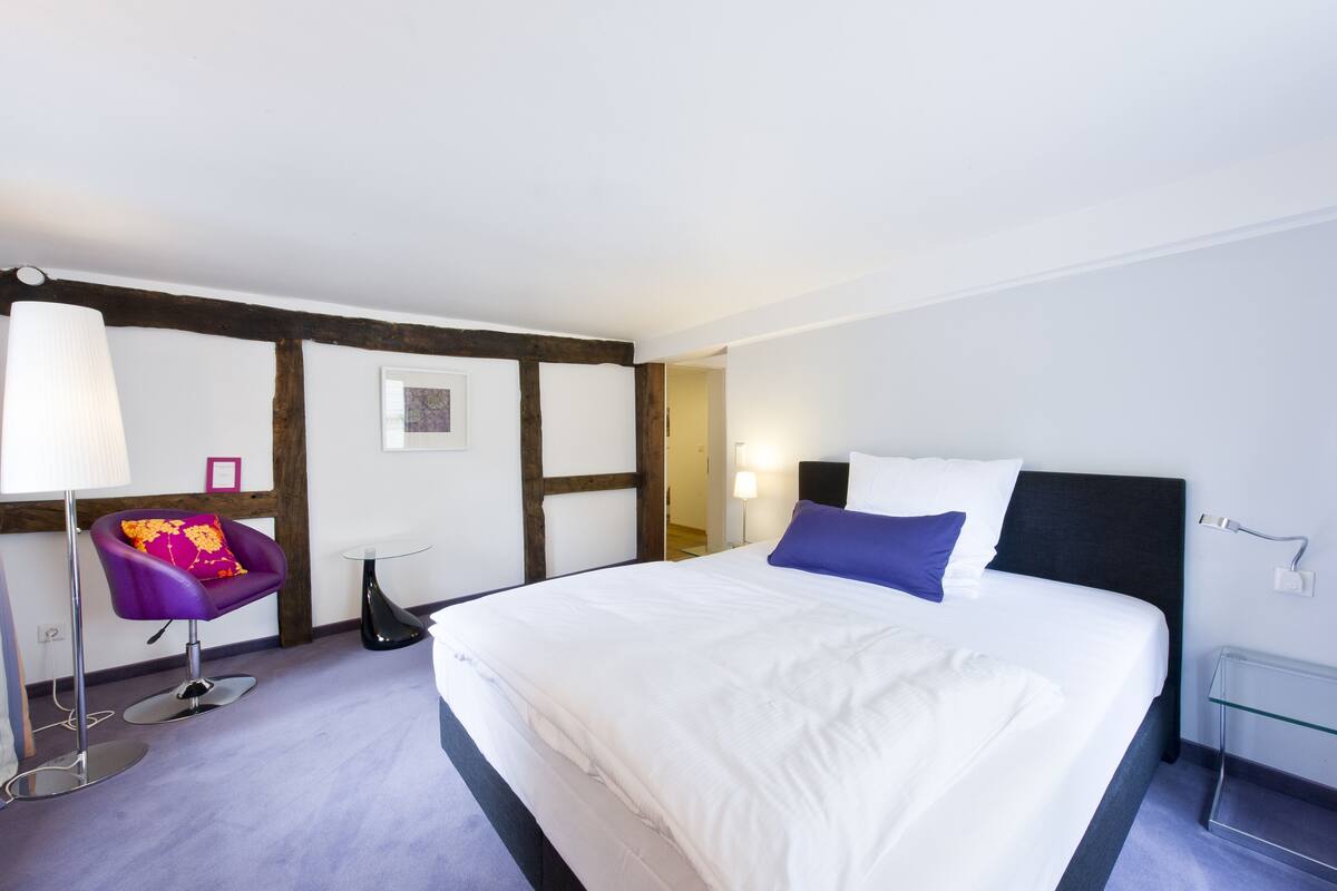 A modern hotel room with a large bed, a chair, a bedside table, and a lamp.