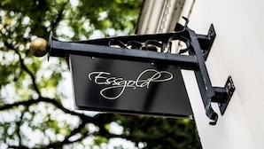 A black metal sign with the word "Essgold" in cursive script, mounted on a building with a white wall and a tree in the background. - Essgold (Ratingen)