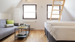 A small, well-lit room with a wooden floor, a grey sofa, a small table with books, a bed with white bedding, and a wooden ladder. - Essgold (Ratingen)