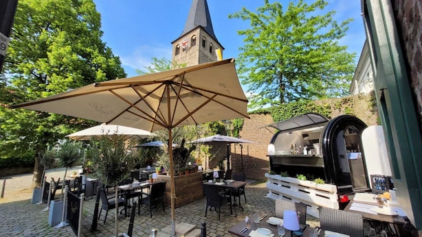 An outdoor patio with tables and umbrellas, a brick wall, and a tower in the background. - Essgold (Ratingen)