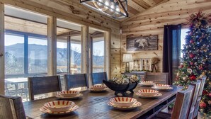 Comedor de madera con una mesa grande dispuesta para seis personas, un árbol de Navidad y vistas a la montaña a través de amplios ventanales.