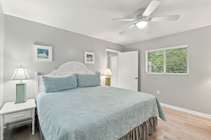 A bedroom with a bed, two bedside tables, a ceiling fan, and a window with a view of greenery. - Beach-Inspired Naples Park Home | Heated Pool (Naples)