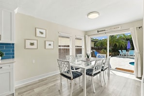 A dining area with a glass table and white chairs, a kitchen with white cabinets, and a sign reading 'Lifes a Beach'. - Beach-Inspired Naples Park Home | Heated Pool (Naples)