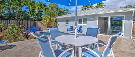 A patio with a round table and blue chairs, a blue umbrella, and a pool area.