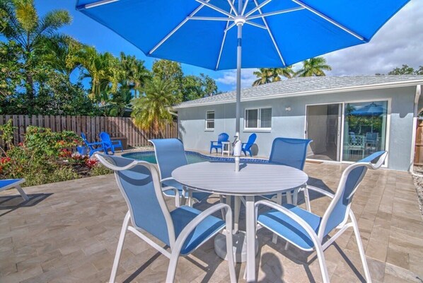 A patio with a round table and blue chairs, a blue umbrella, and a pool area.