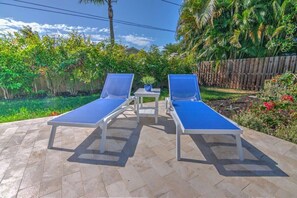 Deux chaises longues dotées de coussins bleus, sur une terrasse carrelée, entourée d’une végétation luxuriante et d’une clôture en bois.