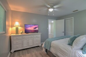 Un dormitorio con cama, televisor sobre una cómoda, ventilador de techo y puerta que da a otra habitación.