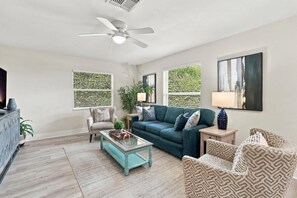 Un salon avec un canapé de couleur turquoise, des fauteuils à motifs, une table basse et un ventilateur de plafond.