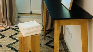 A room with a view of greenery through a glass door, a black desk with a framed picture, and a small wooden stool with folded towels.