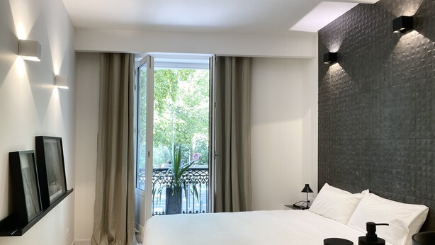A modern hotel room with a large bed, a wooden side table, a black lamp, and a view of a balcony with greenery outside.