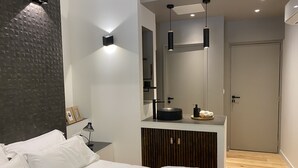A modern bedroom with a bed, bedside table, lamp, and a bathroom vanity with a mirror.