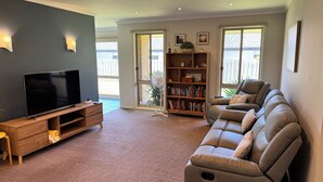 A living room with a flat-screen TV on a wooden stand, a beige sofa, a bookshelf, and a sliding glass door leading to a balcony. - Shore to Impress (Inverloch)