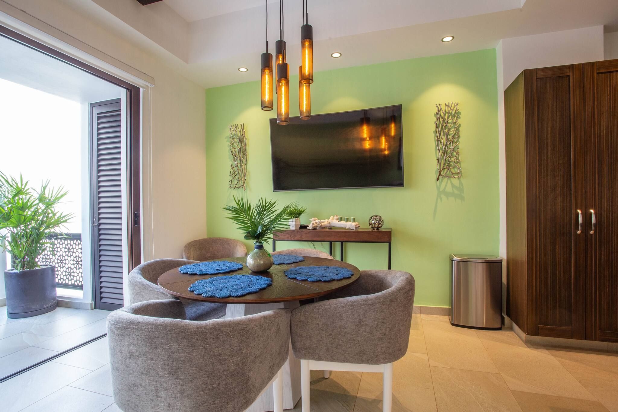 A modern living room with a round table, two grey chairs, a flat-screen TV mounted on the wall, and a potted plant.
