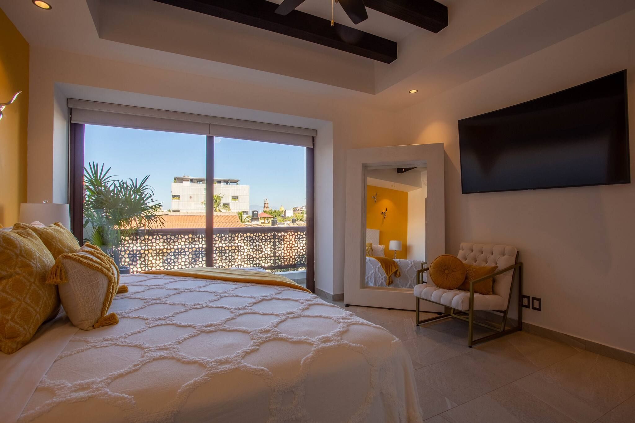 A bedroom with a large bed, a chair, a TV, and a balcony with a view of buildings.