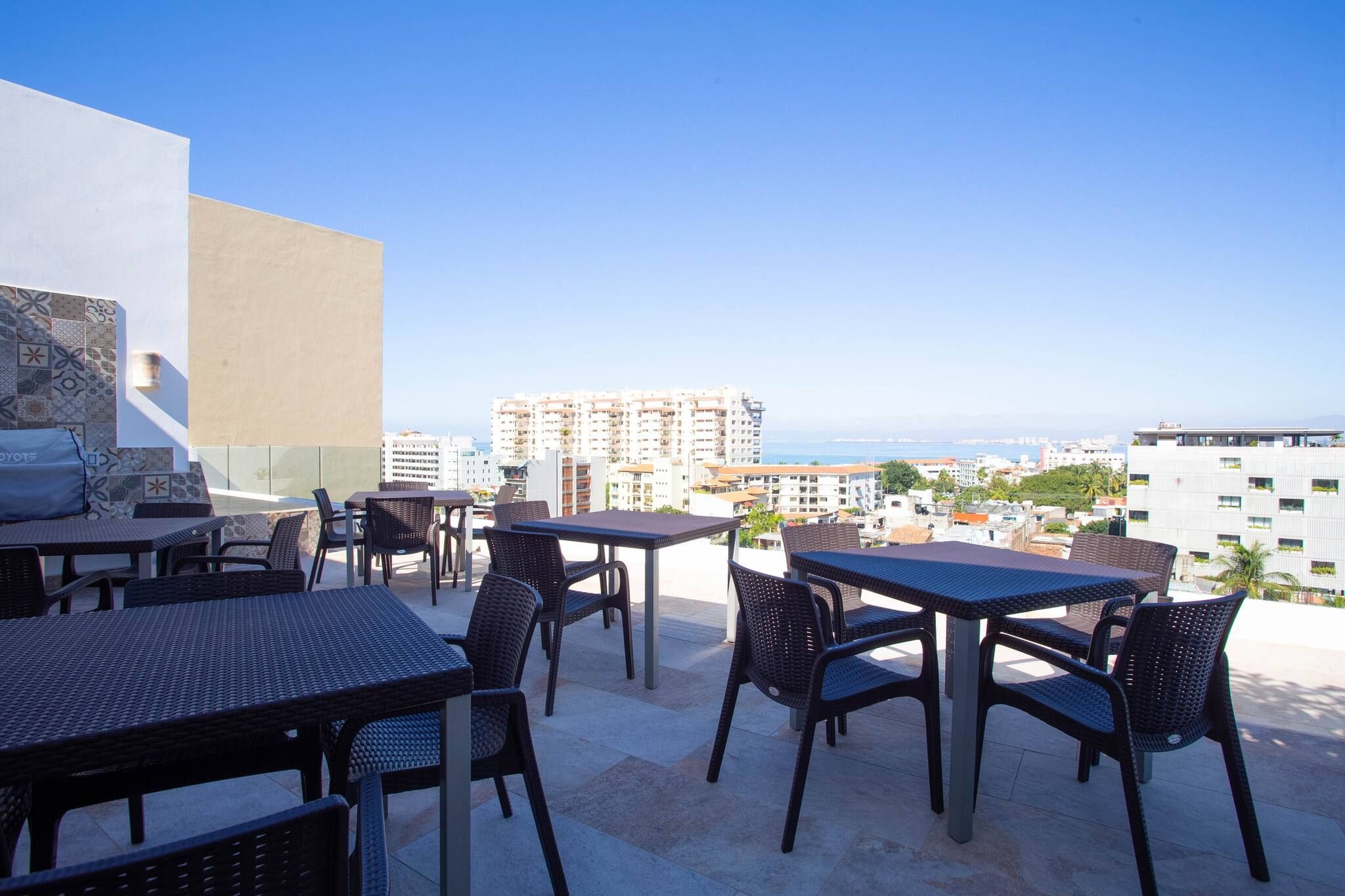 An outdoor terrace with tables and chairs, overlooking a cityscape and the sea.