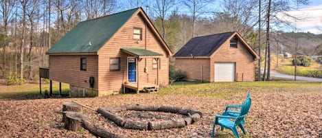 Two wooden cabins with green roofs, a fire pit, and a blue chair in a leaf-covered clearing.