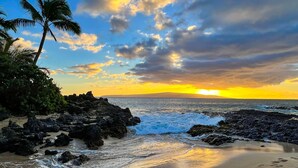 Une plage bordée de rochers, avec des palmiers, et un coucher de soleil sur l’océan.