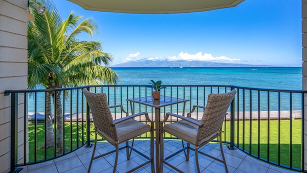 Un balcon avec deux fauteuils en osier et une table, d’où l’on peut admirer une mer d’un bleu profond et une chaîne de montagnes lointaine.