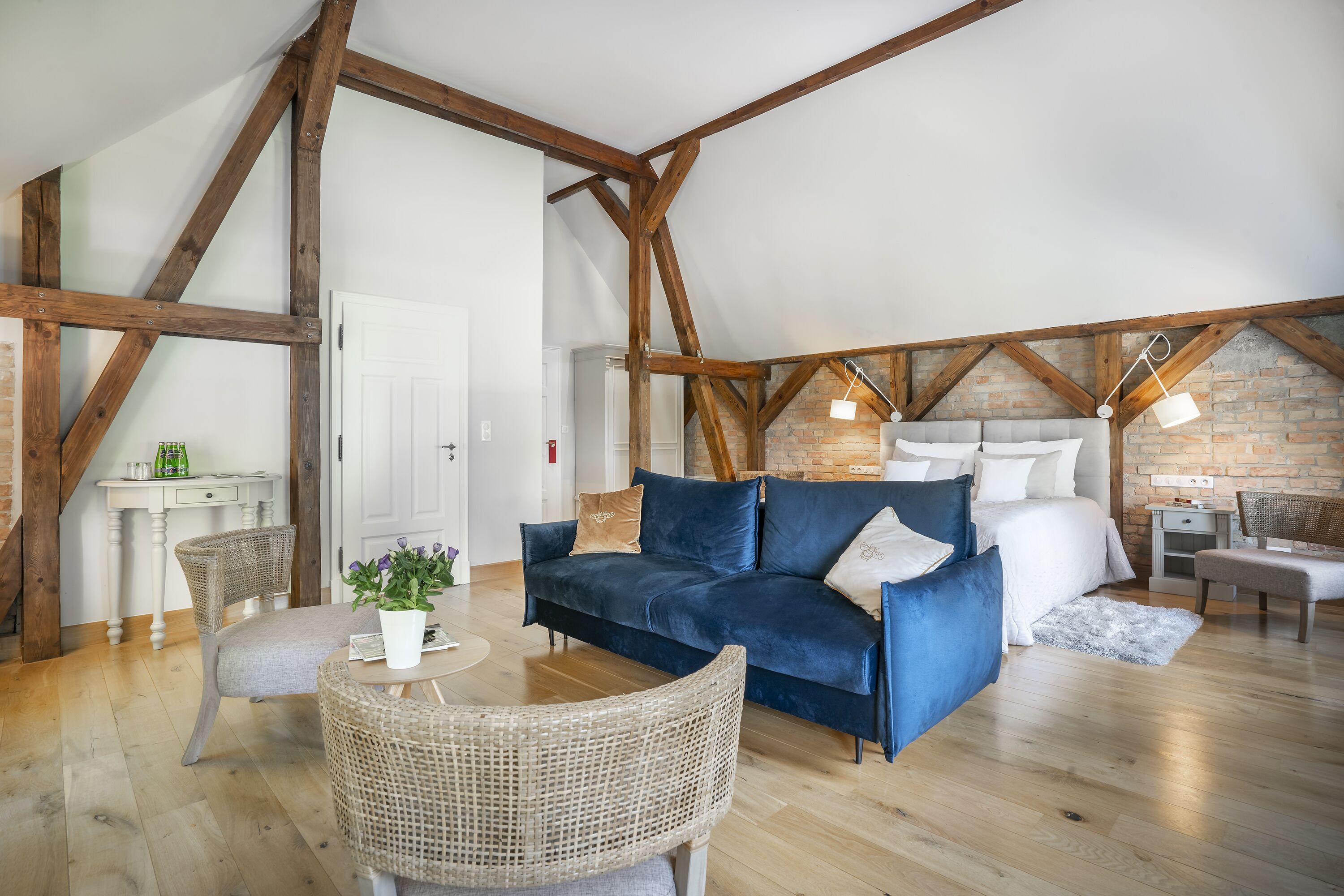 Un cuarto estilo loft con vigas de madera a la vista, un sofá de terciopelo azul, un sillón de mimbre y una pequeña mesa con una planta.