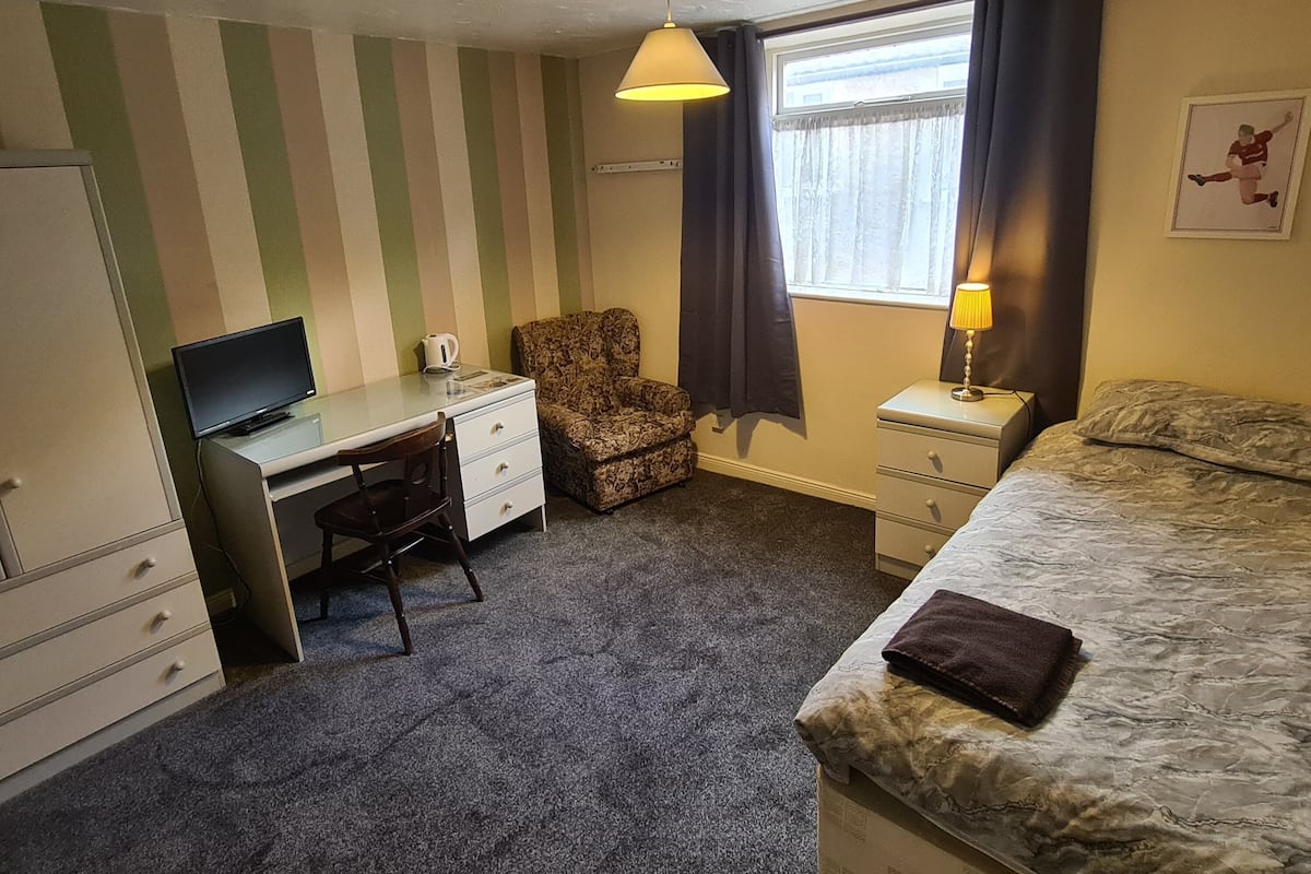 A compact hotel room with a single bed, a desk with a chair, a small table with a lamp, and a window with curtains.